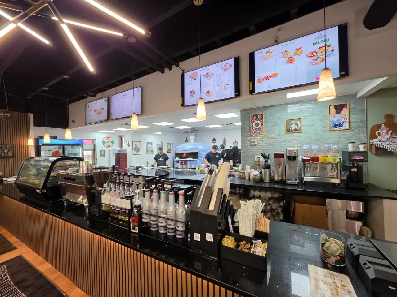 The finished Al Manakeesh service counter: espresso bar, backlit menu boards, a tiled wood-fired oven, and pendant lighting over black stone.