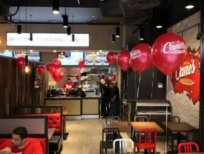 A completed Raising Cane’s dining room: an illuminated welcome sign over the order counter, a painted brick feature wall, and red banquette and chair seating.